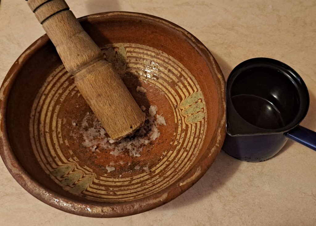 Clay bowl with crushed garlic and pestle, small cezve of oil beside it.