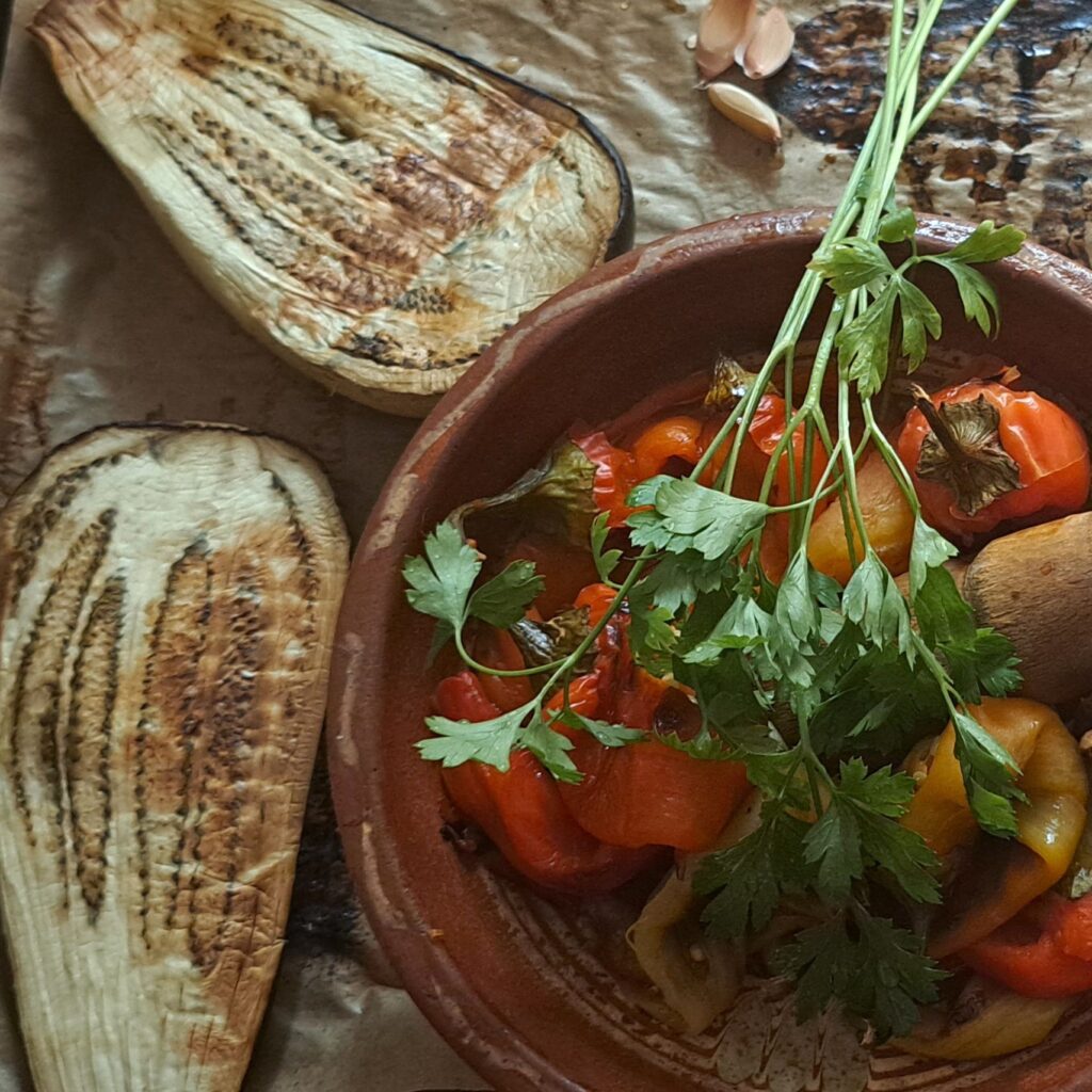 Roasted peppers in clay plate, halved roasted eggplant, whole garlic cloves, and fresh parsley.