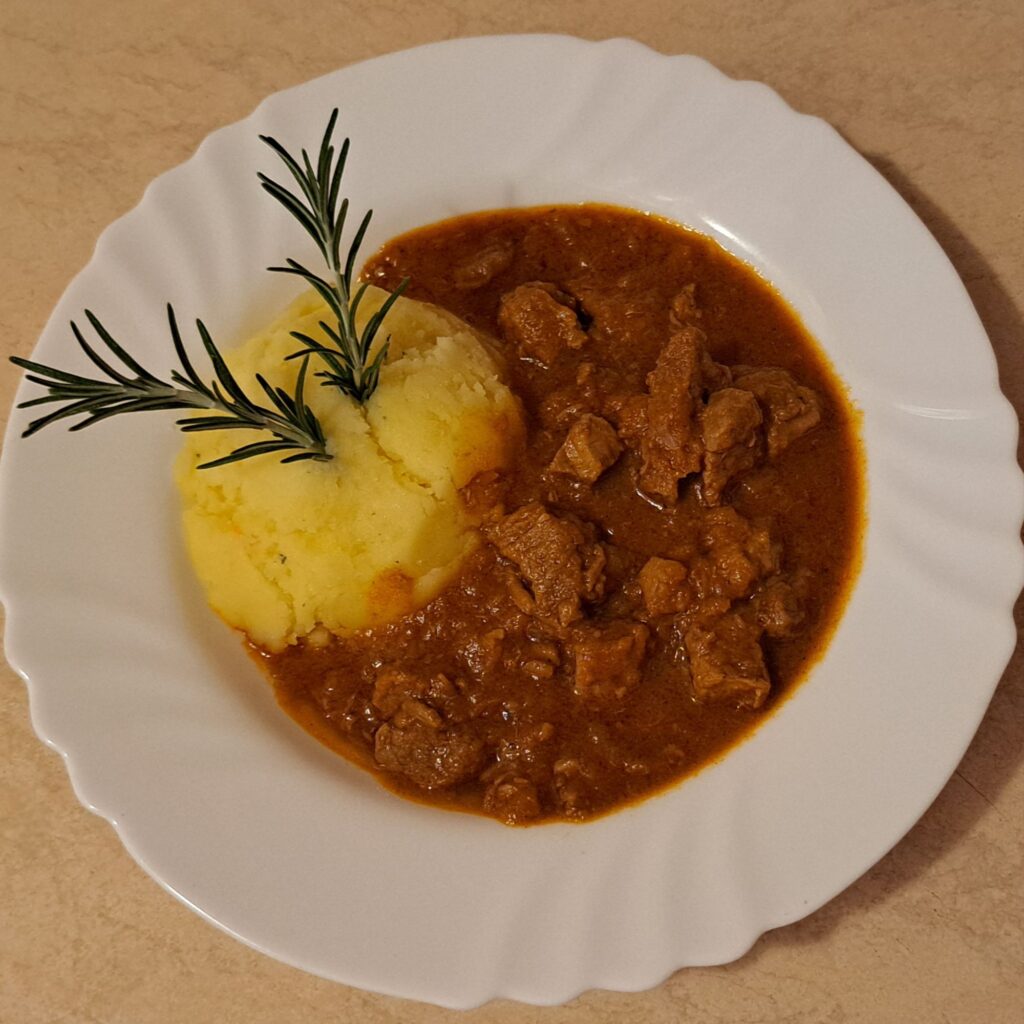 Plate of Homemade Beef Goulash served with mashed potatoes and decorated with two rosemary sprigs.
