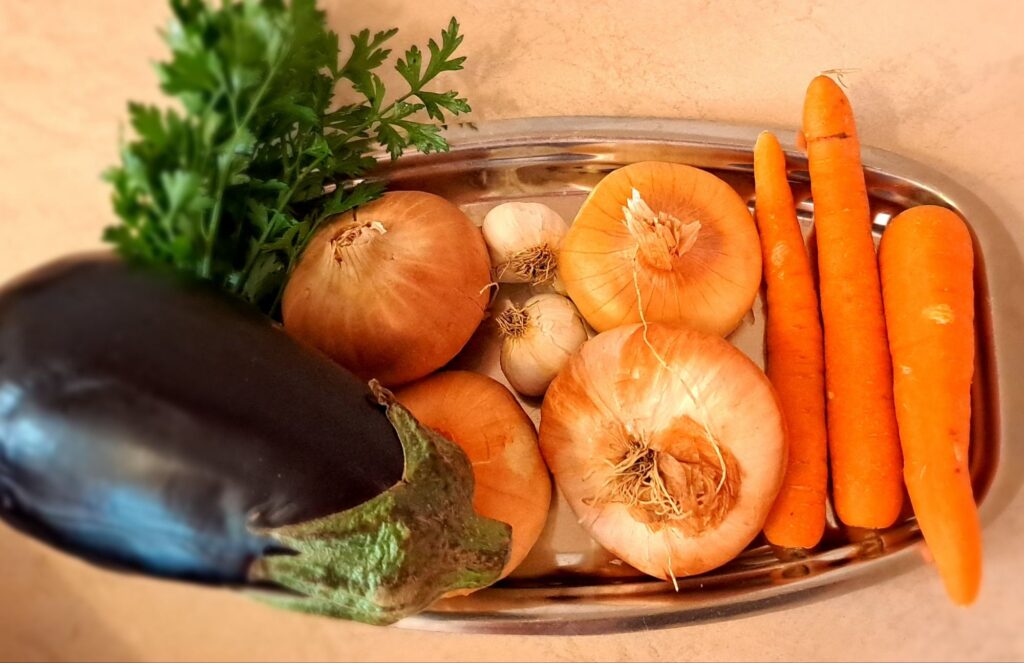Onions, garlic cloves, carrots, eggplant, parsley placed whole on kitchen table.