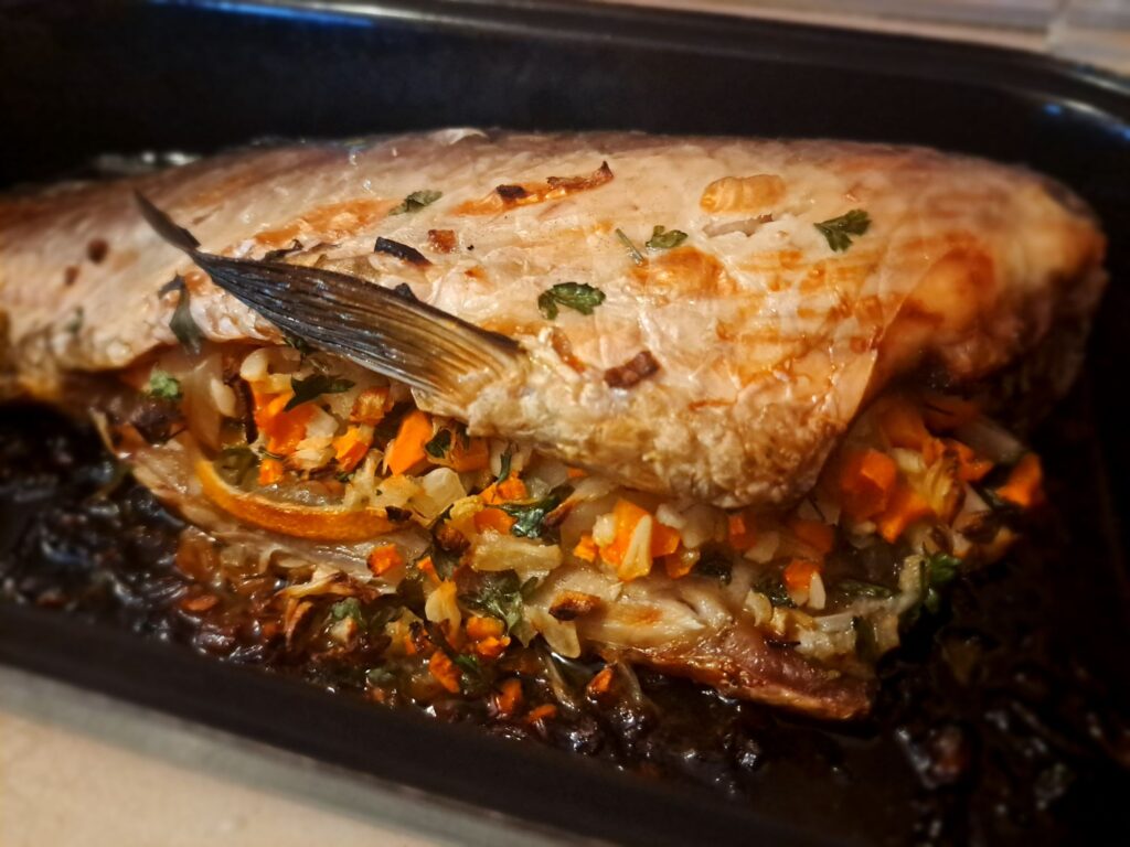 Whole stuffed carp after baking, surrounded by roasted onions, garlic, carrots, eggplant, parsley, and lemon slices directly in the tray.