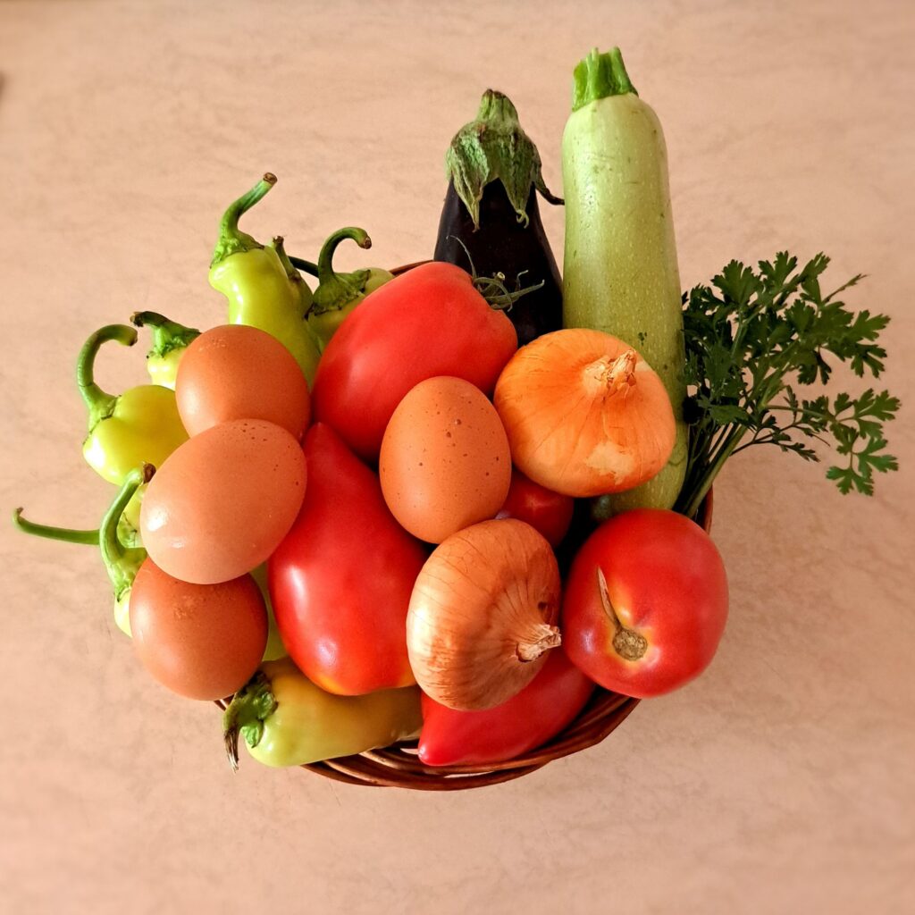 Whole onions, peppers, tomatoes, zucchini, eggplant, parsley, and eggs arranged in a woven wooden basket.
