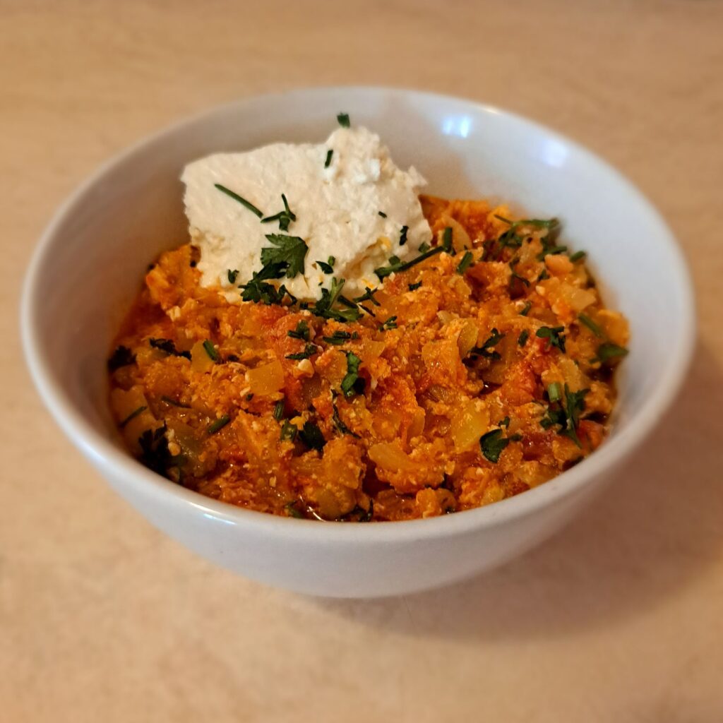 Fully cooked vegetable and egg mixture served in a deep bowl, garnished with a piece of sheep cheese and chopped parsley.