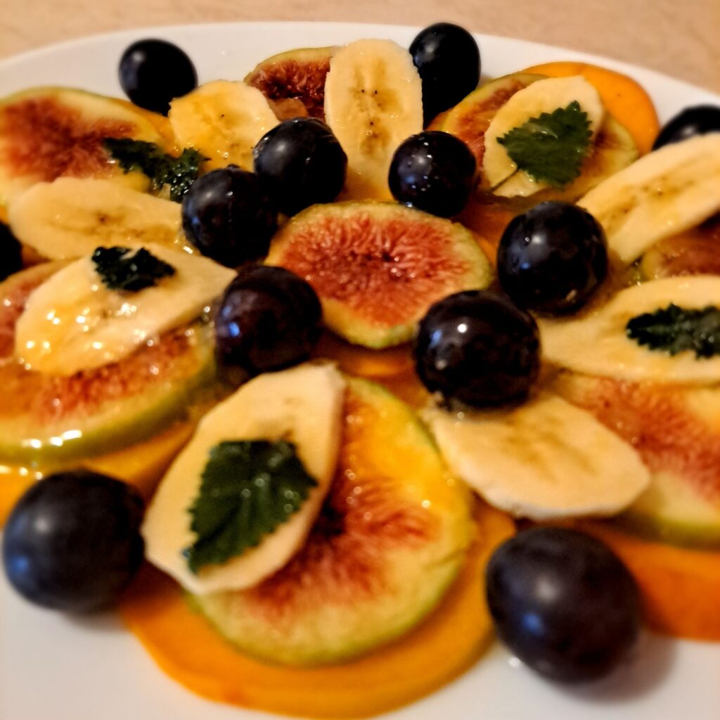 Side angle of fruit carpaccio with thin peach, fig, and banana slices layered with grapes, mint, honey, and orange juice.
