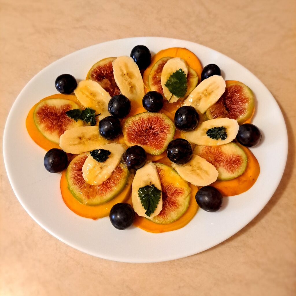 Thin slices of peach, fig, and banana arranged in circles with grapes, mint leaves, honey, and orange juice on top.