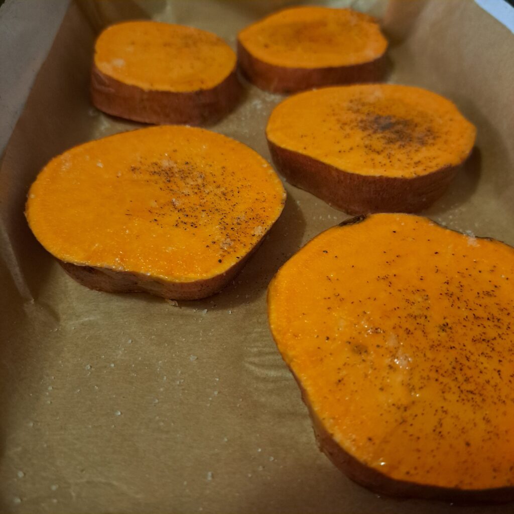 Unpeeled sweet potato slices on baking paper in a tray before roasting.