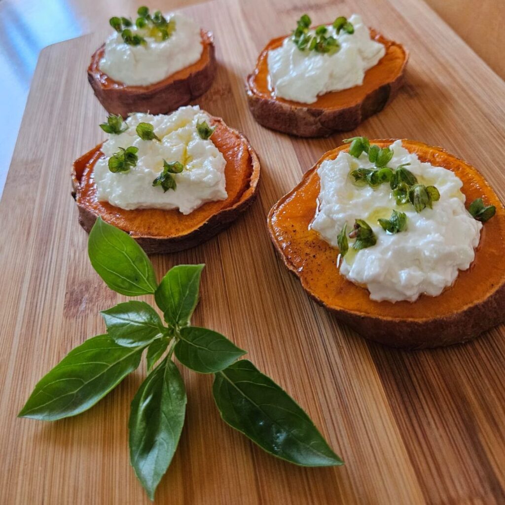 Sweet potato bites with ricotta and microgreens on a wooden board with a basil sprig.