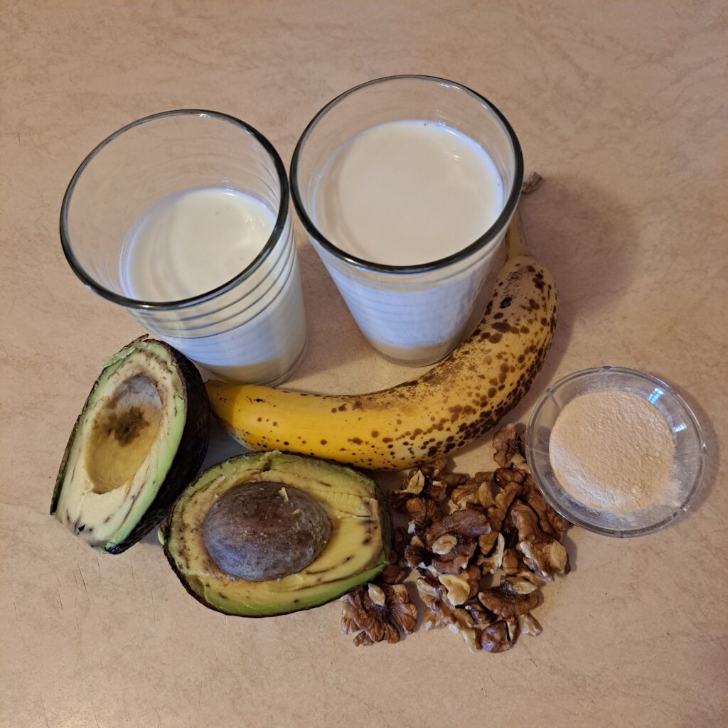 All ingredients for Baobab Avocado Banana Smoothie arranged on a kitchen table: halved avocado, banana, baobab powder, a glass of milk, a glass of yogurt, and walnut pieces.