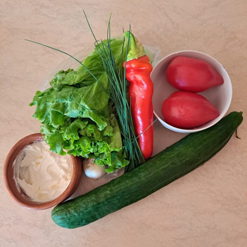 Whole lettuce, cucumber, tomatoes, garlic, yogurt, fresh chives, and red bell pepper displayed as salad ingredients.