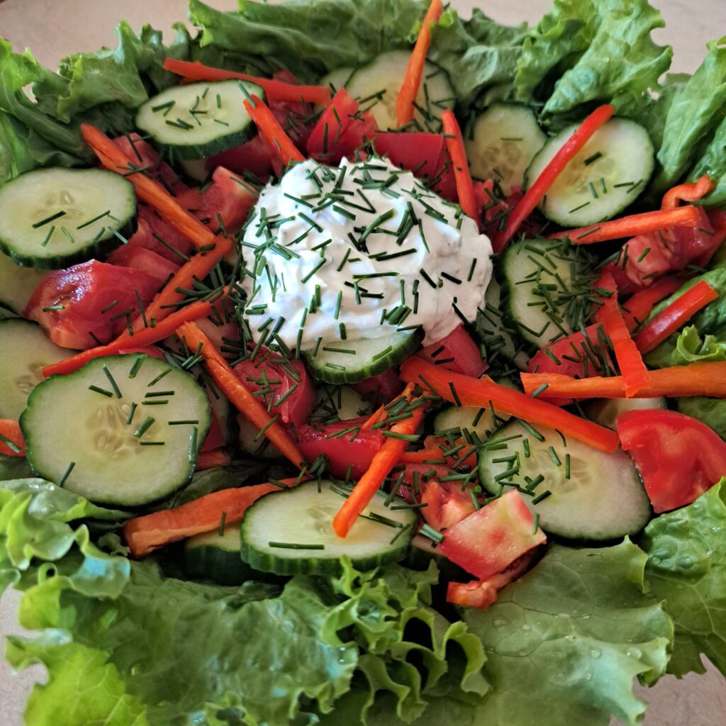 Close-up view of garden salad with creamy yogurt dressing showing lettuce, cucumber, tomatoes, chives, and bell pepper.