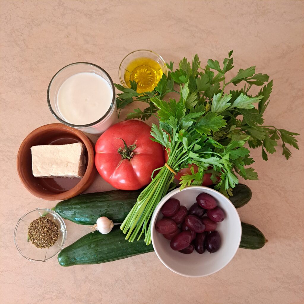 Whole cucumbers, tomatoes, garlic, yogurt, white cheese, black olives, fresh parsley, olive oil, and oregano on a kitchen counter.