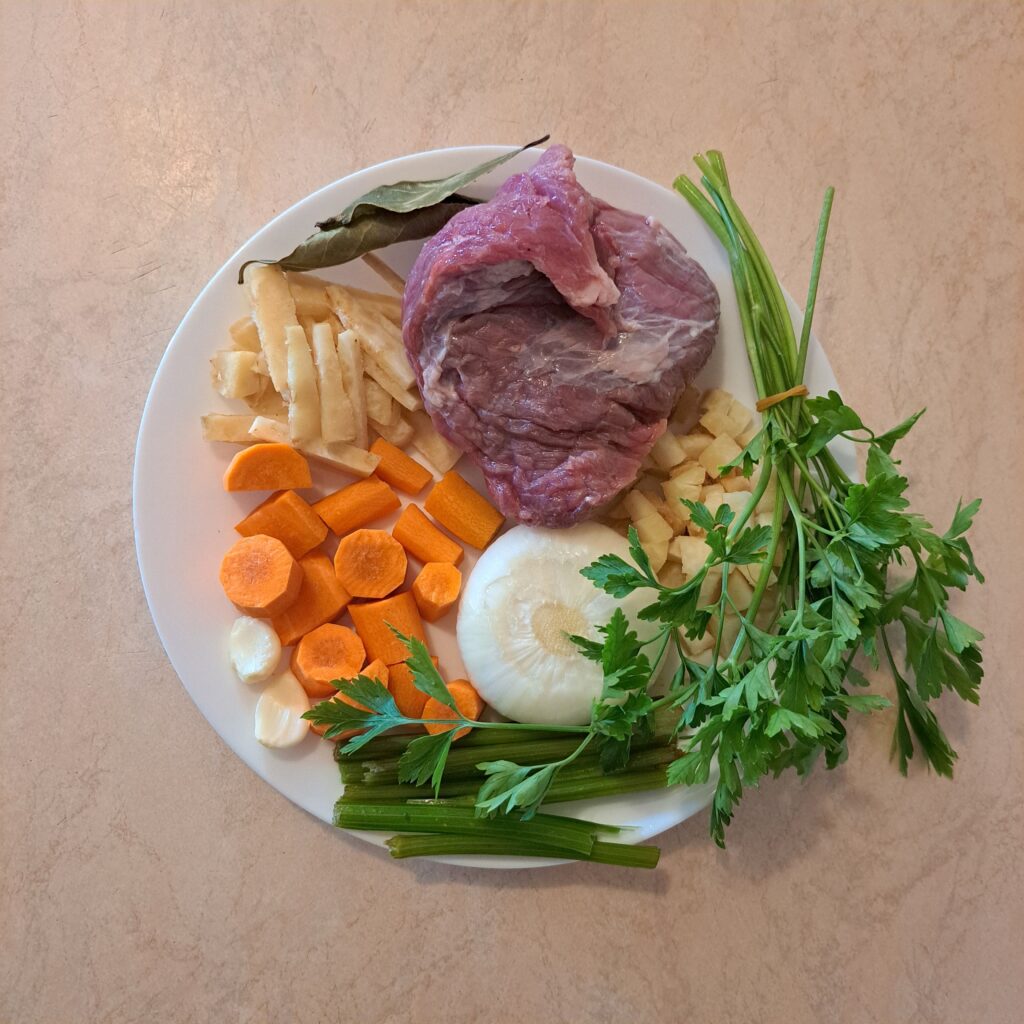 Whole beef, chopped parsnip, chopped celery root, celery stalks, large carrot pieces, parsley bunch, whole peeled onion, bay leaf, and peeled garlic cloves arranged on a white plate.