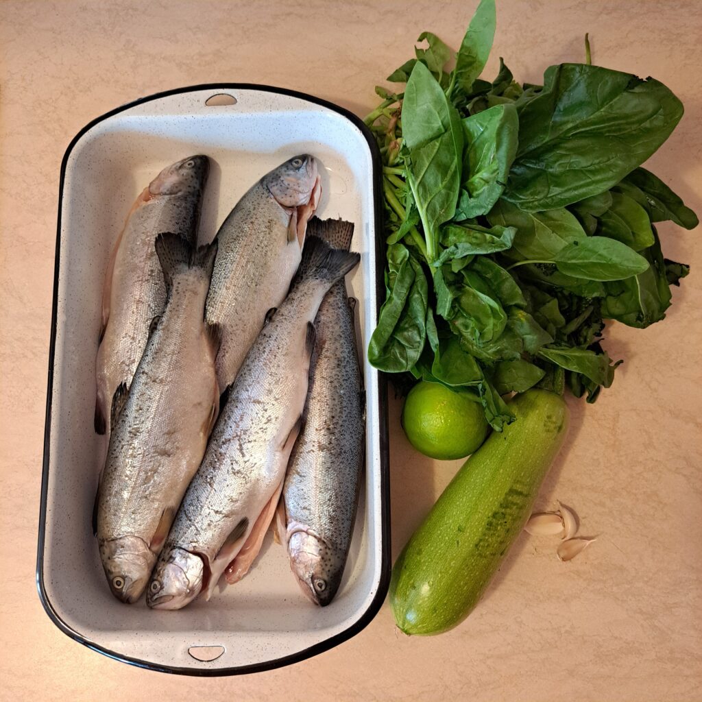Five cleaned Californian trout in a baking tray, spinach, zucchini, lime, and garlic cloves.