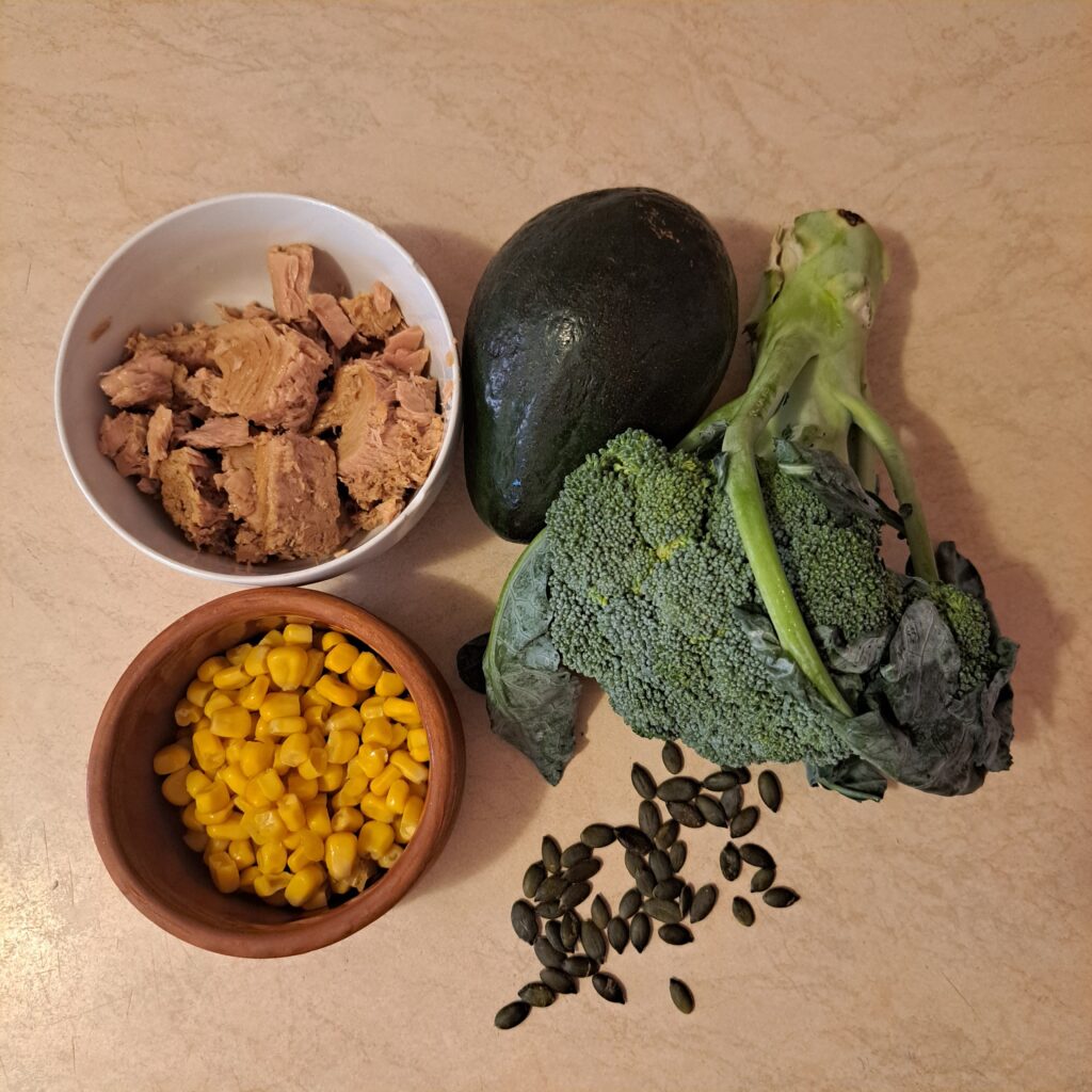 Ingredients for Avocado Broccoli Tuna Salad: whole broccoli, avocado, drained canned tuna, corn, and pumpkin seeds on kitchen counter.