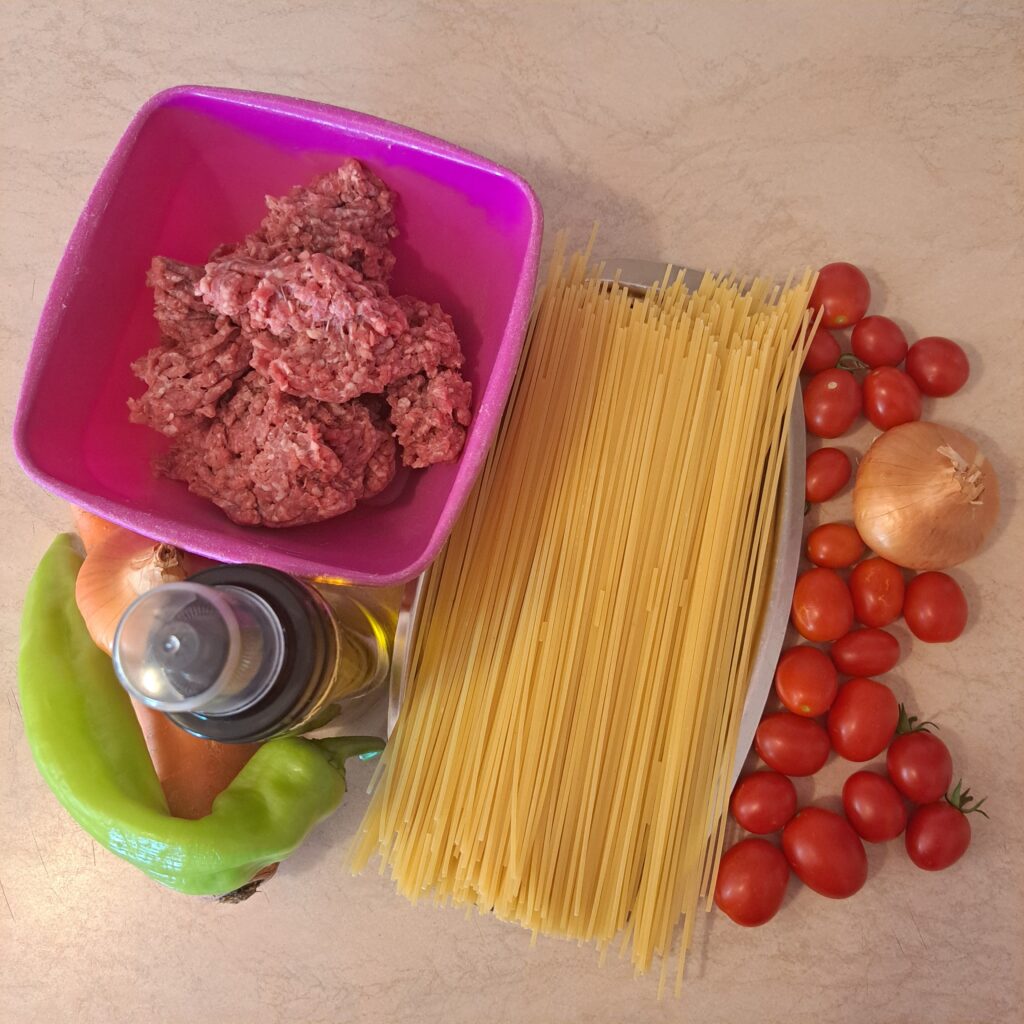 Ingredients for Homemade Beef Vegetable Spaghetti arranged on a kitchen countertop, including spaghetti, ground beef, cherry tomatoes, onion, carrot, bell pepper, and cooking oil.