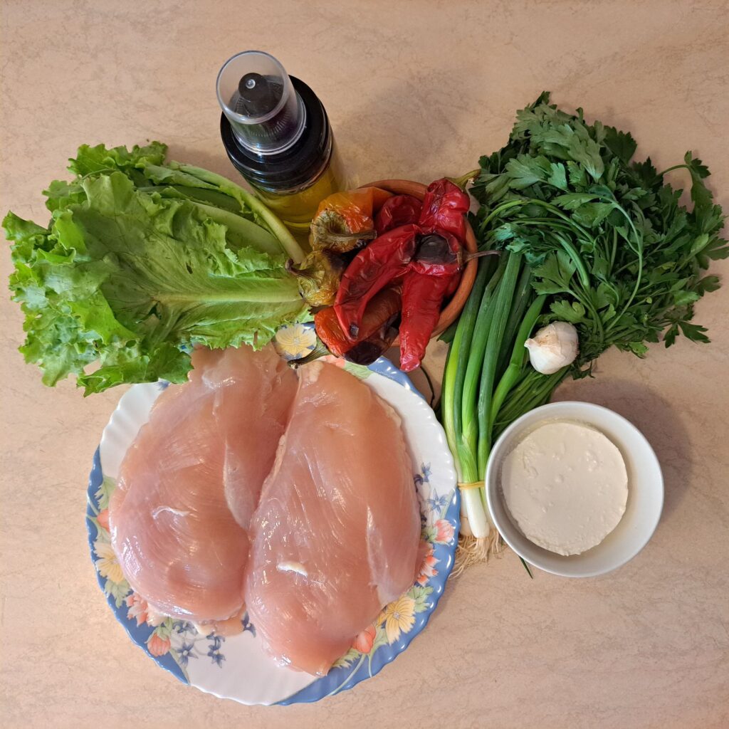 All ingredients for Mini Rolled Chicken Bites laid out on a table, including chicken steaks, cream cheese, red peppers, spring onion, parsley, arugula, and garlic.