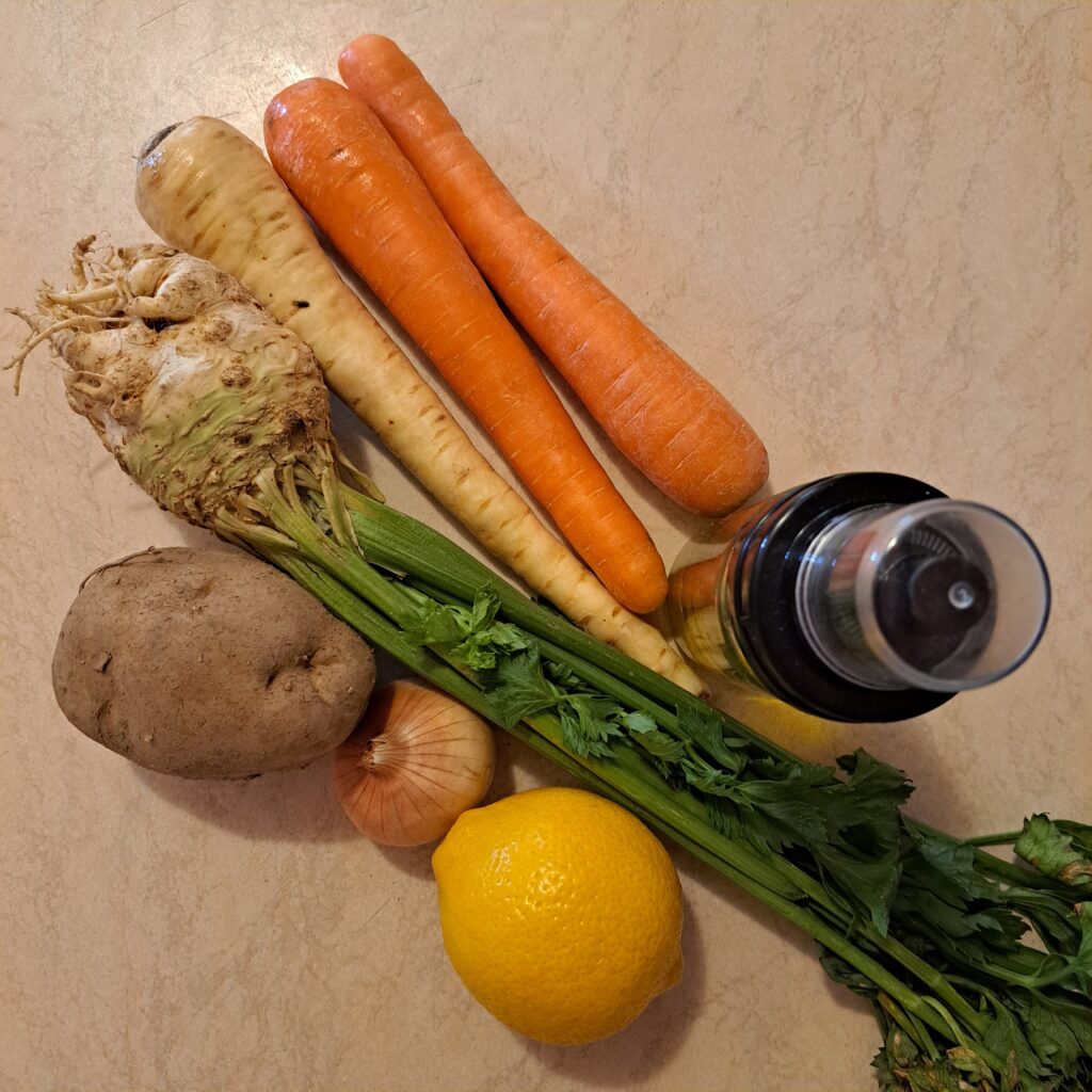 Carrots, celery, parsnip, potato, onion, lemon, and oil arranged on a kitchen table.