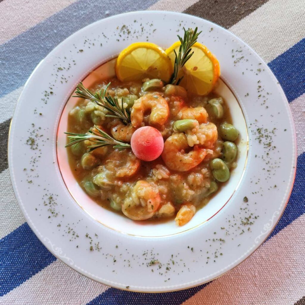 Overhead view of a plate with creamy shrimp broad bean risotto, garnished with rosemary, lemon, and cherry tomato.
