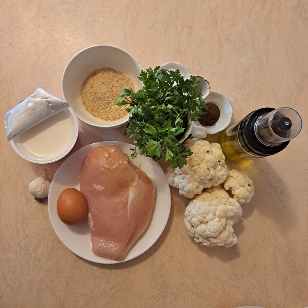 Kitchen table with chicken breast, cauliflower, egg, breadcrumbs, garlic, parsley, salt, and pepper.