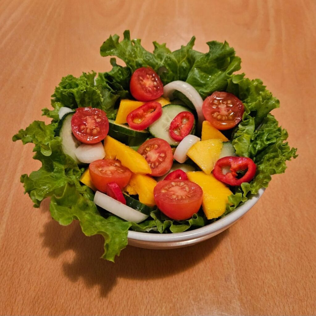 Overhead view of mango cucumber garden salad in a bowl, with colorful layers of mango, cucumber, tomatoes, onion, and bell pepper on greens.