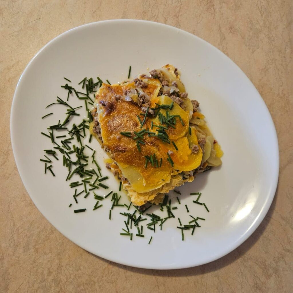 Top-down view of a plated portion of potato zucchini meat moussaka garnished with fresh chives.