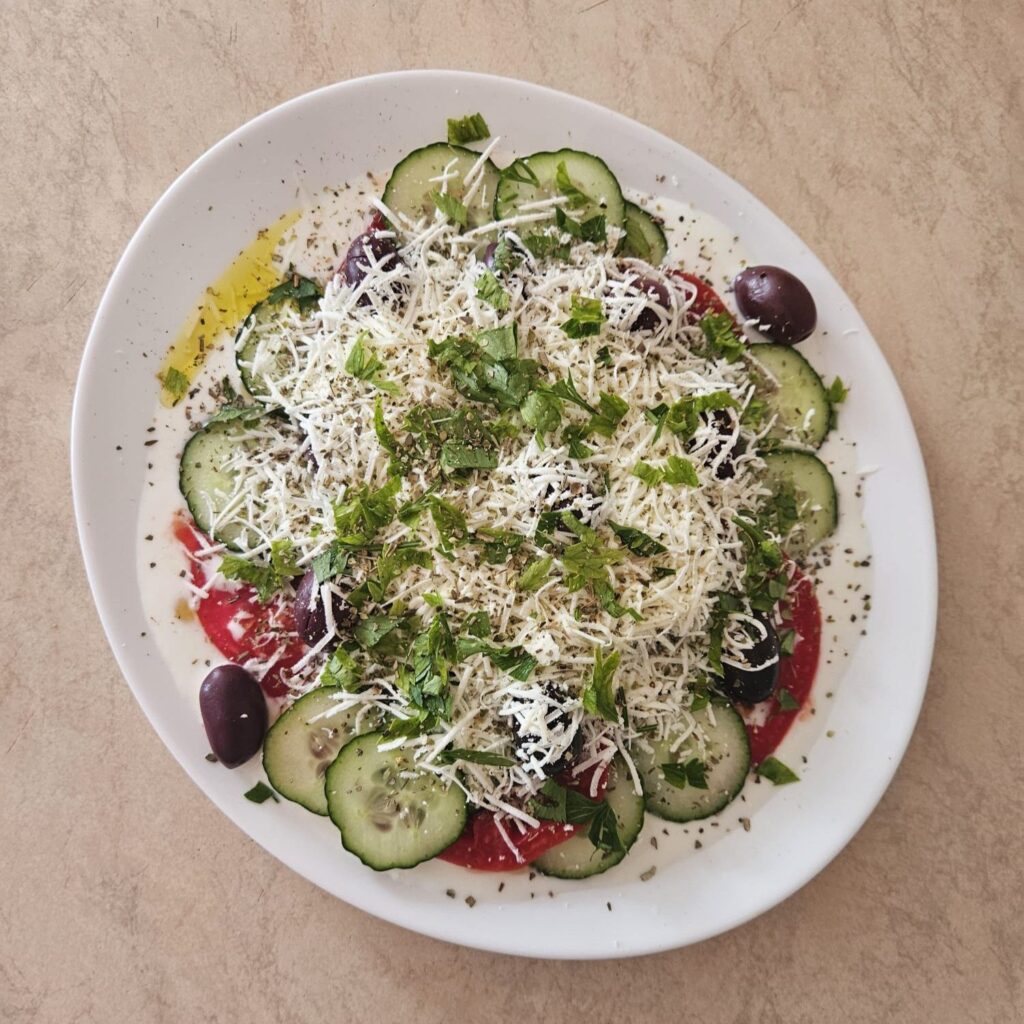 Completed salad with grated white cheese, fresh parsley, and a sprinkle of oregano on top of sliced cucumbers, tomatoes, and black olives.
