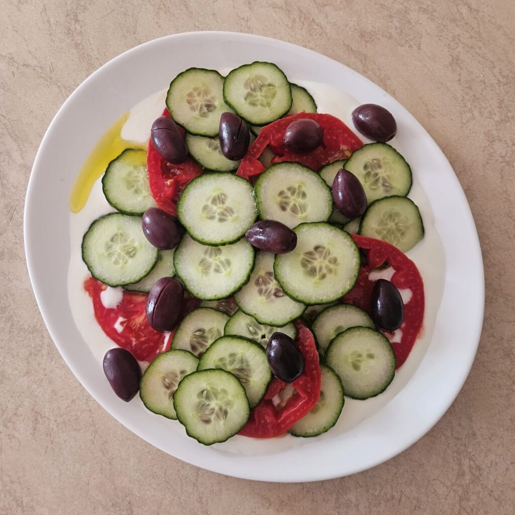 Yogurt and olive oil mixture spread on a plate, topped with sliced cucumbers, tomatoes, and whole black olives.