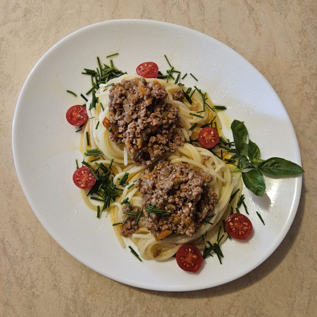 Top-down view of a plate with Homemade Beef Vegetable Spaghetti, topped with beef and vegetable sauce, garnished with chives, basil, and halved cherry tomatoes.