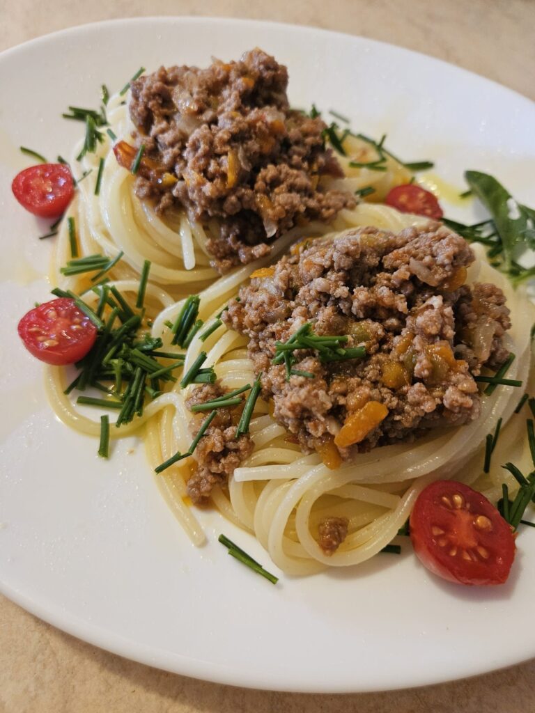 Close-up of a plate with homemade spaghetti topped with beef and vegetable sauce, garnished with chives, basil, and halved cherry tomatoes.