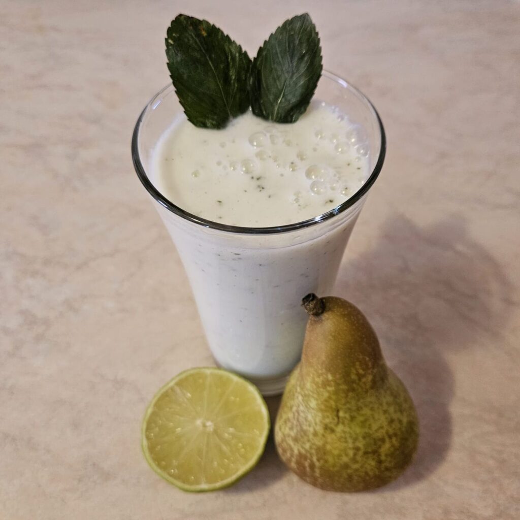 Pear ginger lime smoothie with mint served in a tall glass with pear and half lime.