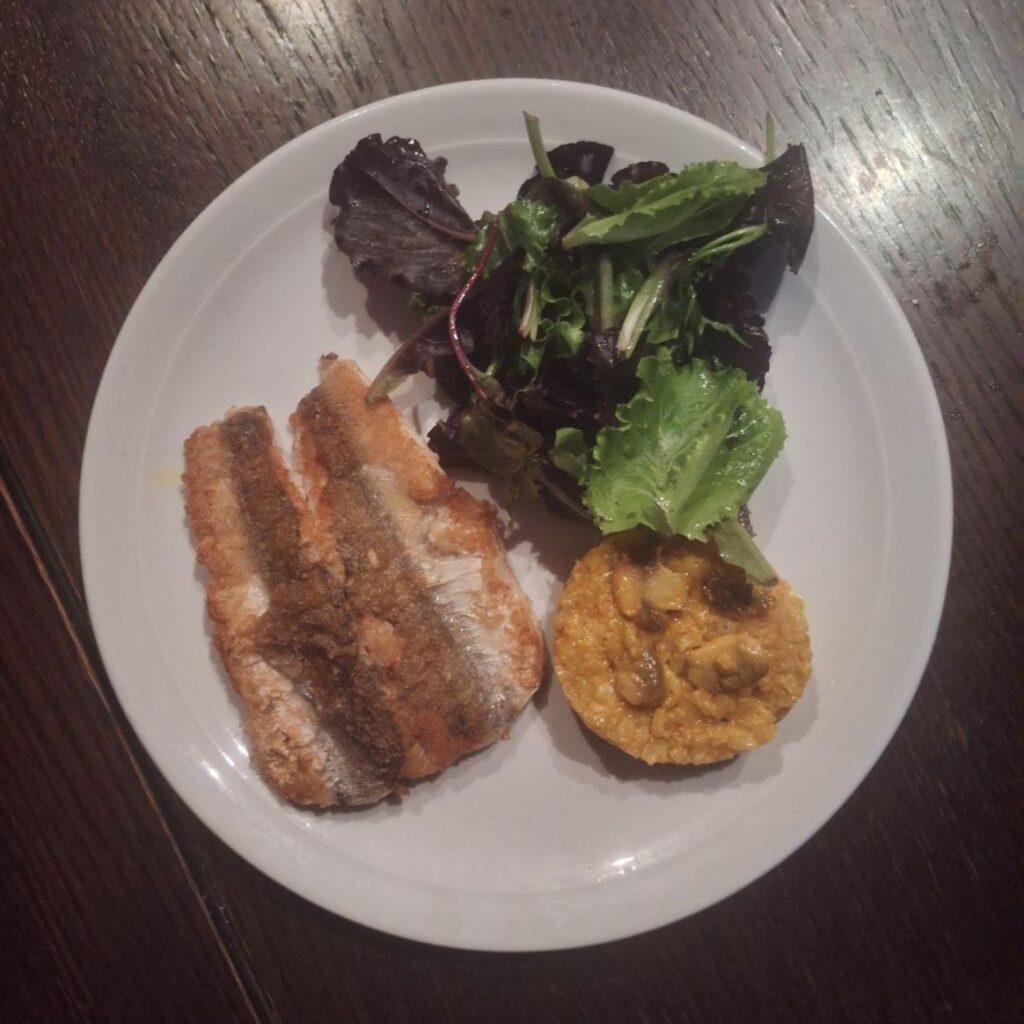 A plate with baked hake fillet, mushroom rice ball, and fresh baby salad.