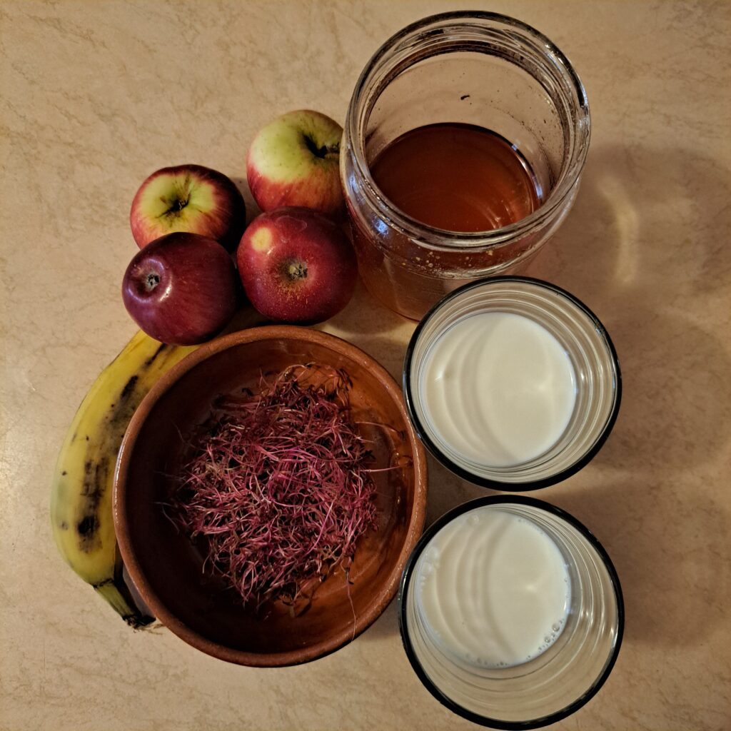 Fresh ingredients for Apple Banana Amaranth Smoothie arranged on a table: whole apples, banana, milk, yogurt, and a handful of amaranth microgreens.