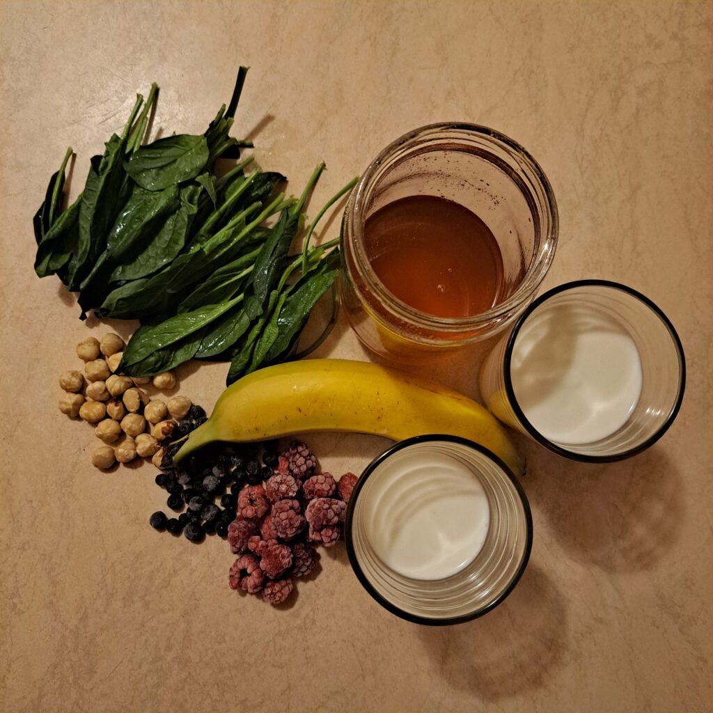 Raspberries, aronia berries, spinach, banana, hazelnuts, yogurt, milk, and honey arranged on a table.