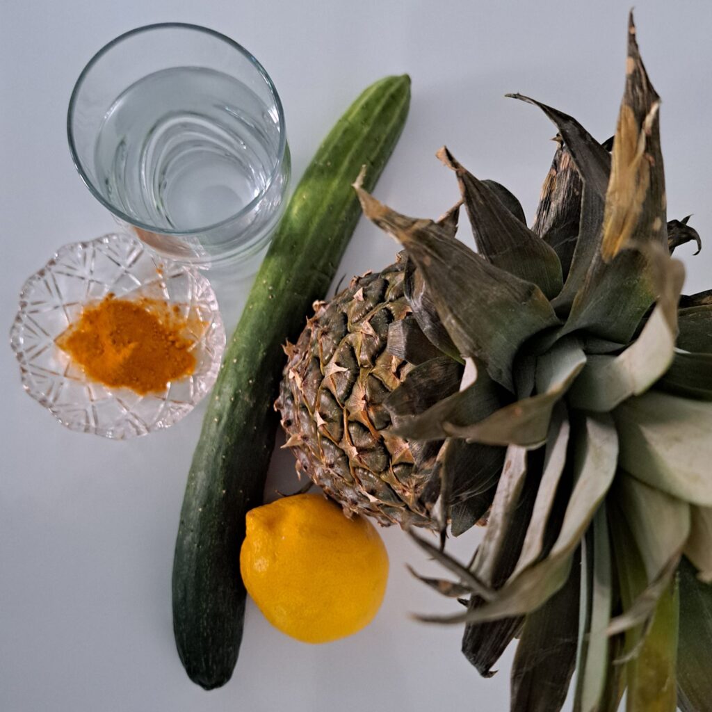 Pineapple, cucumber, turmeric, lemon, and a glass of water arranged on a table.