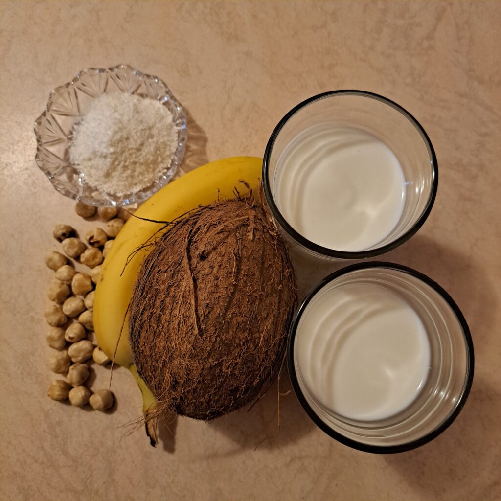 Smoothie ingredients displayed on a table: banana, milk, yogurt, hazelnuts, shredded coconut, and a whole coconut.