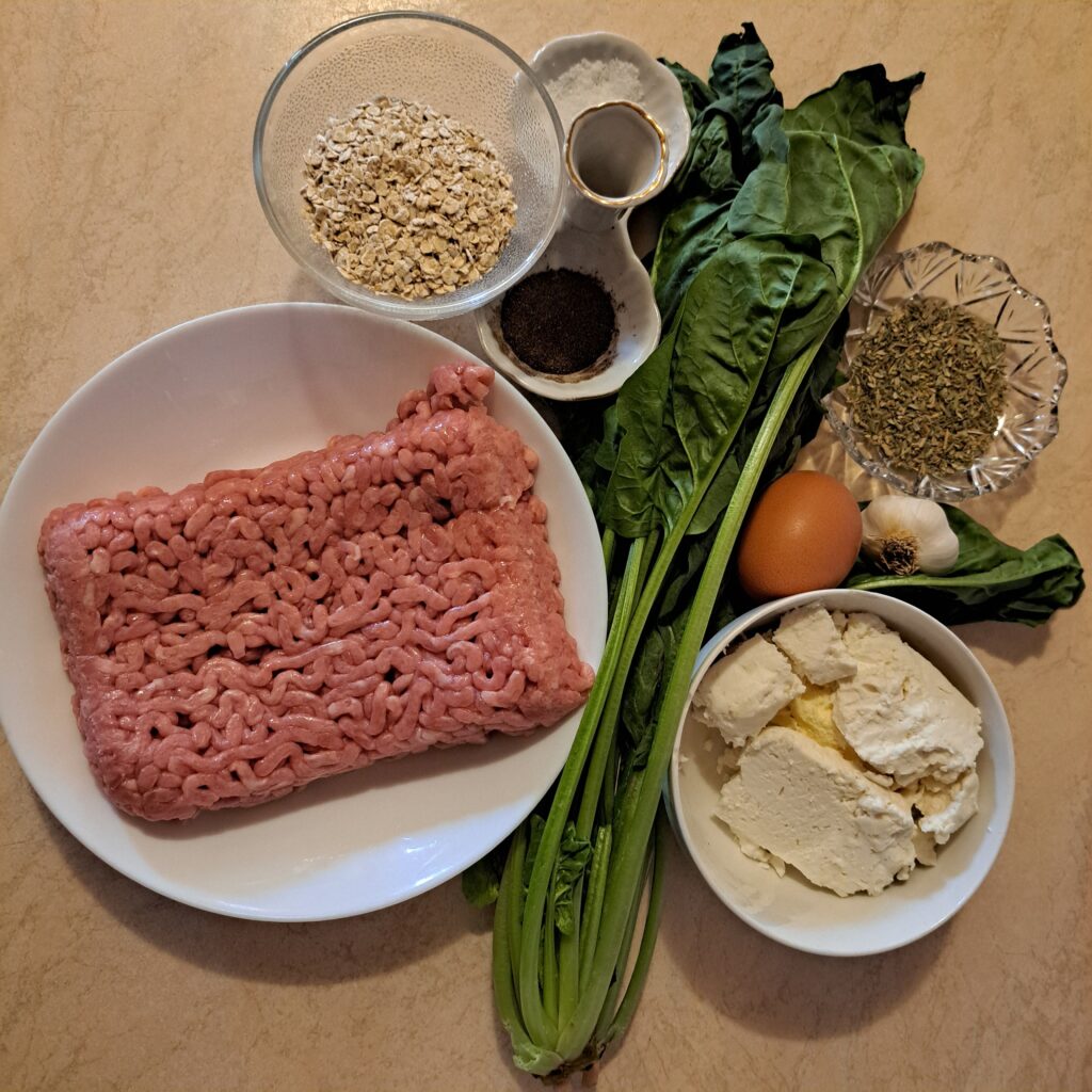 Fresh ingredients for turkey meatballs on a table, including ground turkey, spinach, feta cheese, egg, oats, garlic, dried oregano, salt, and pepper.
