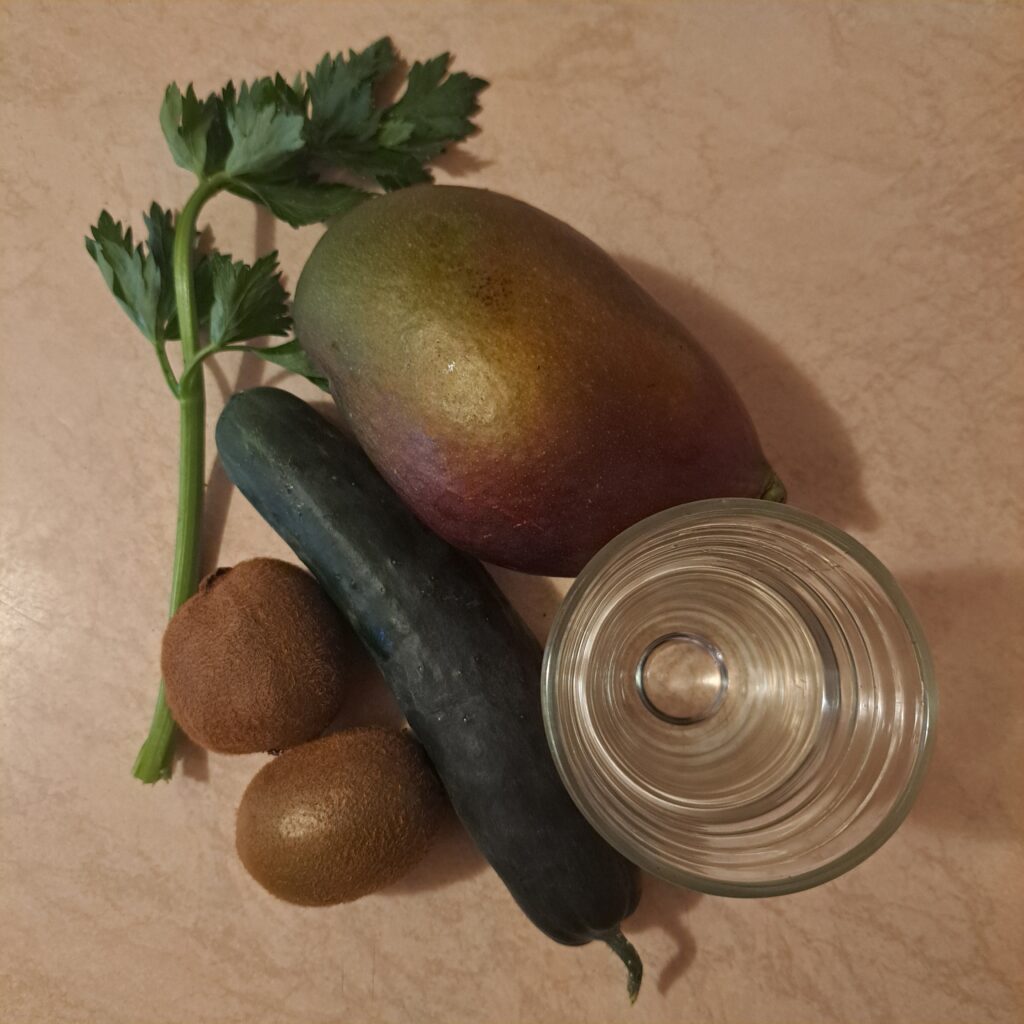 Fresh ingredients for kiwi mango celery juice on a table including mango, kiwi, cucumber, celery stalk, and a glass of water.