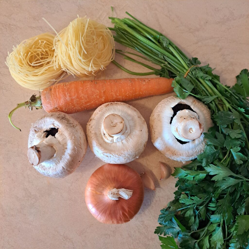 Mushrooms, carrots, onion, garlic, parsley, and thin noodles arranged on a wooden table.