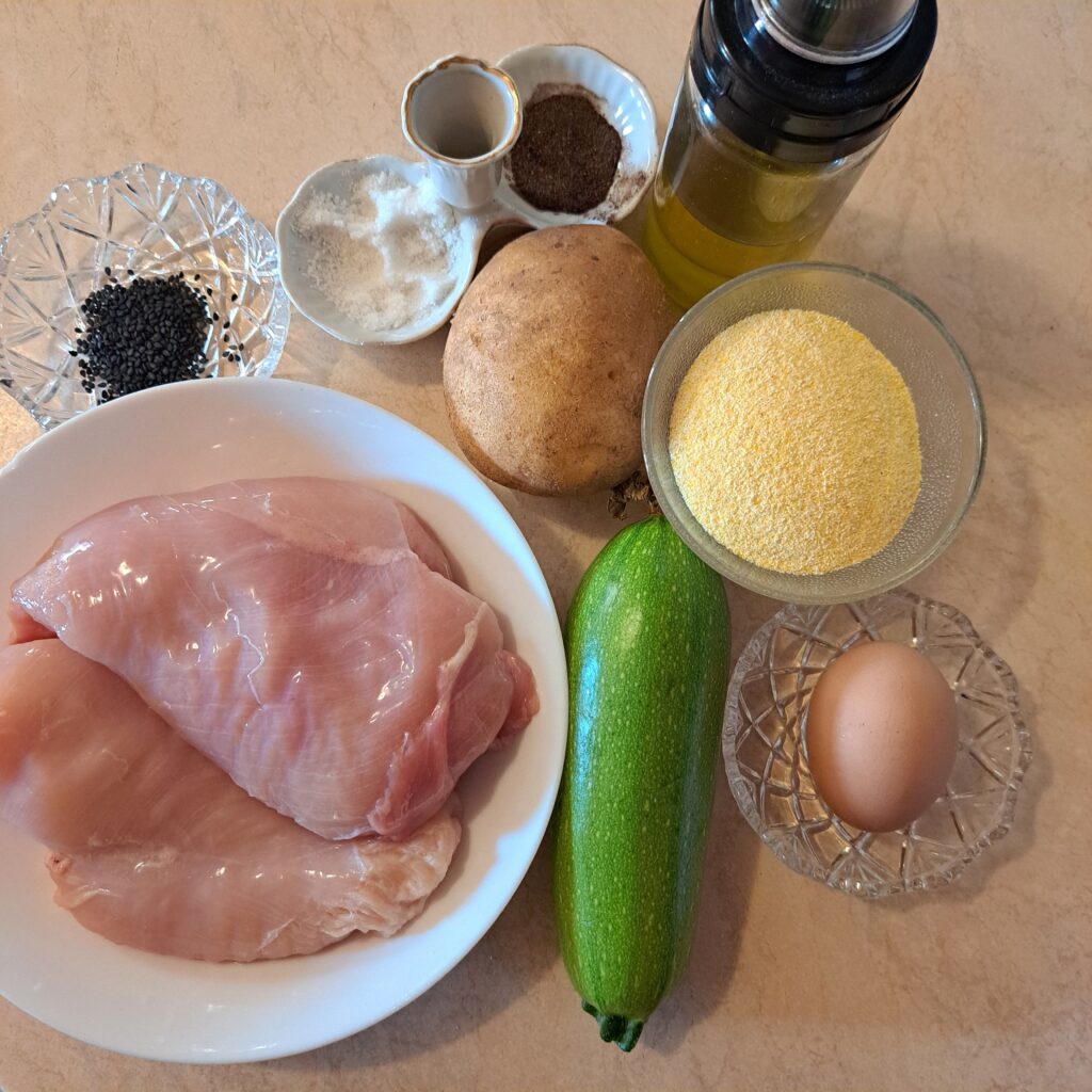 Chicken breast, zucchini, potato, cornmeal, egg, black sesame, olive oil, salt, and pepper on a table.