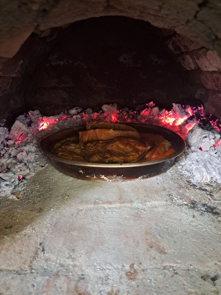 Round baking tray with roasted pork inside a wood fired oven seen from outside.