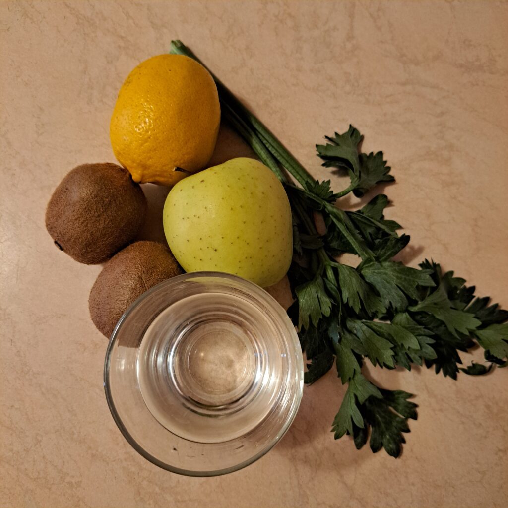 Green apple, two kiwis, lemon, celery stalks, and half a glass of water arranged on a table before making the juice.