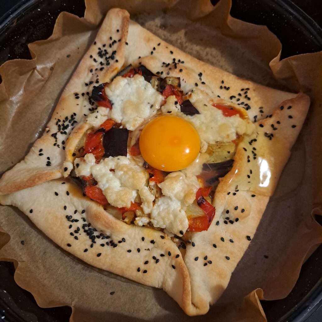Golden-brown galette on parchment paper in a baking tray, topped with a sunny egg, roasted vegetables, and melted cheese.