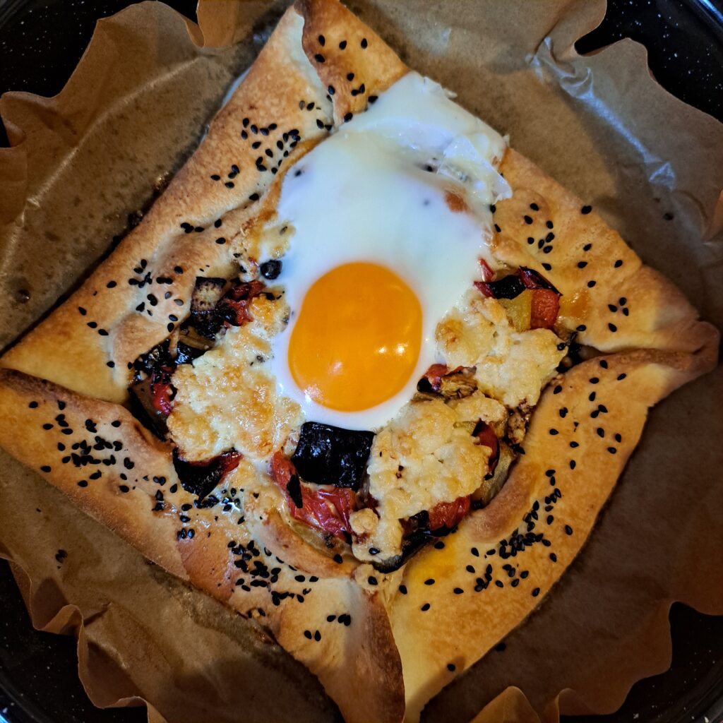 Golden-brown galette on parchment paper in a baking tray, topped with a sunny egg, roasted vegetables, and cheese visible.