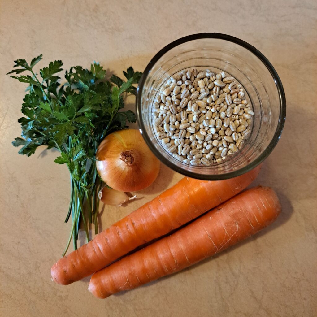 Barley in a glass, carrots, onion, garlic clove, and parsley bunch arranged on a table.