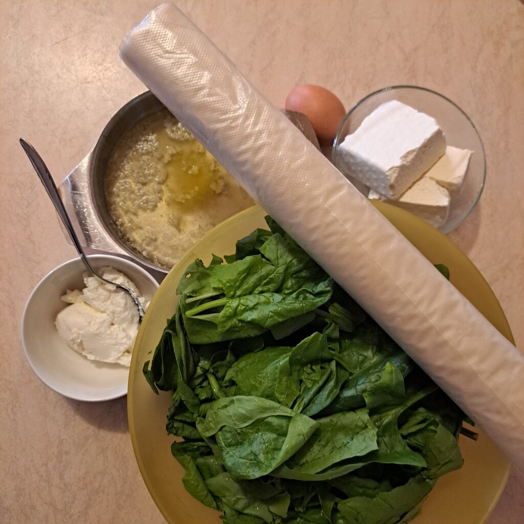 Fresh ingredients arranged neatly on a kitchen counter: spinach, feta cheese, cream cheese, egg, phyllo pastry sheets, and melted butter. 