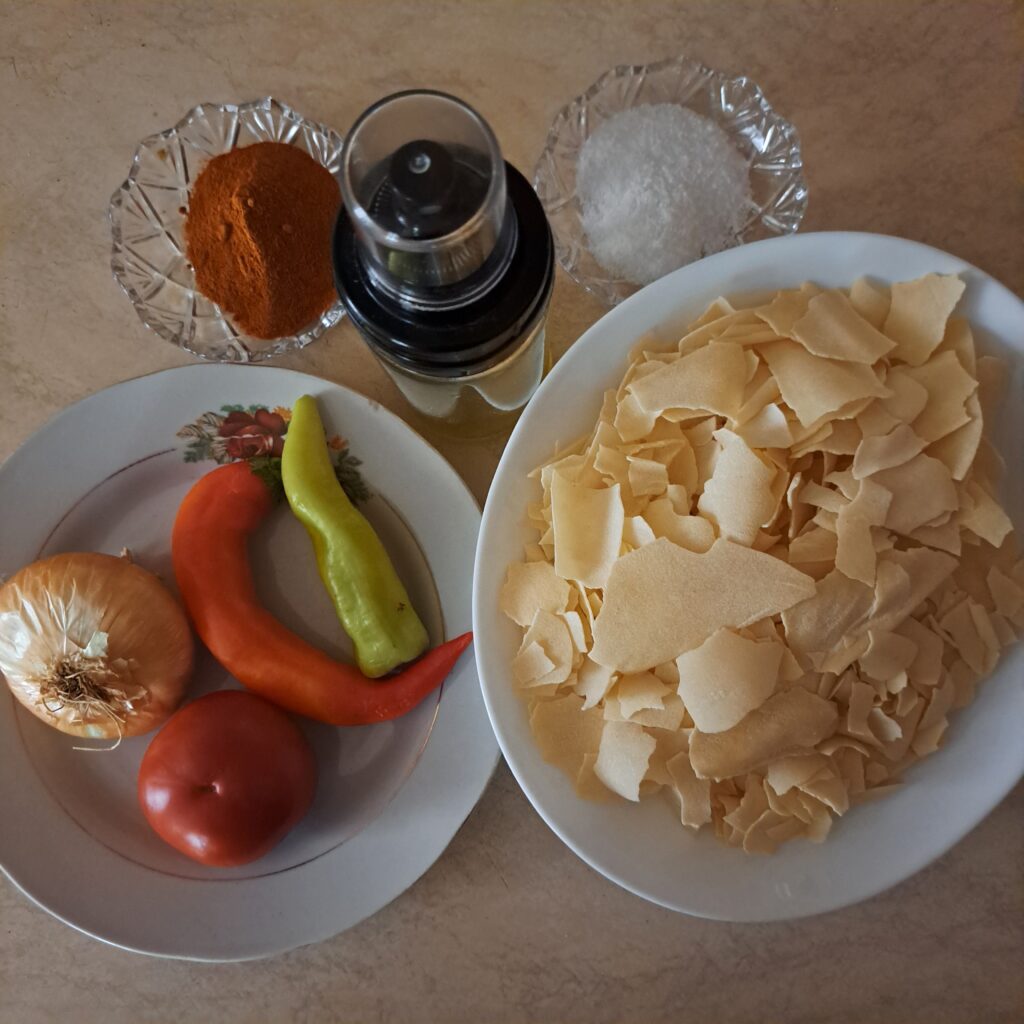 Broken pasta sheets, red and green bell peppers, onion, tomato, salt, ground dried red pepper, and olive oil on a surface.