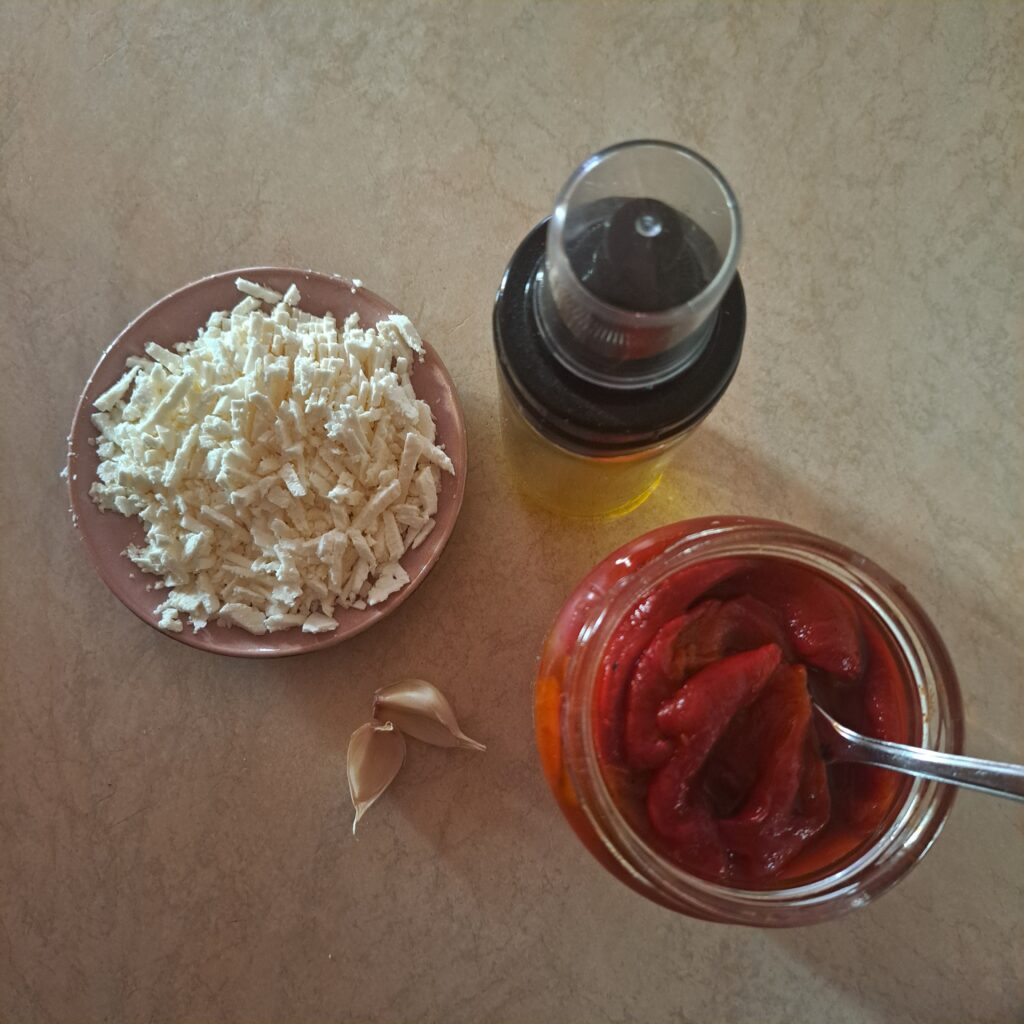 Fermented red peppers in a jar, goat cheese, garlic clove, and olive oil arranged on a kitchen table.