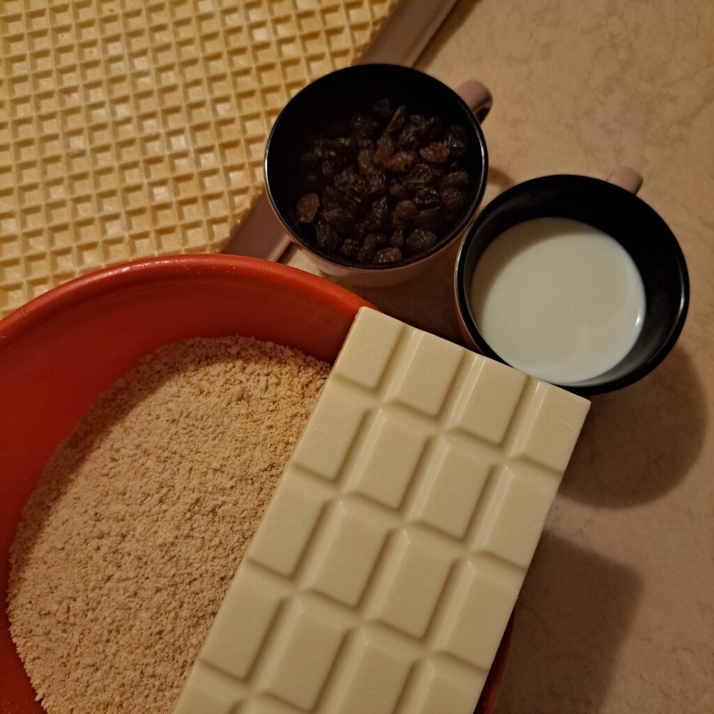 Wafer sheet, crushed biscuits, white chocolate, milk, and raisins on a kitchen table.