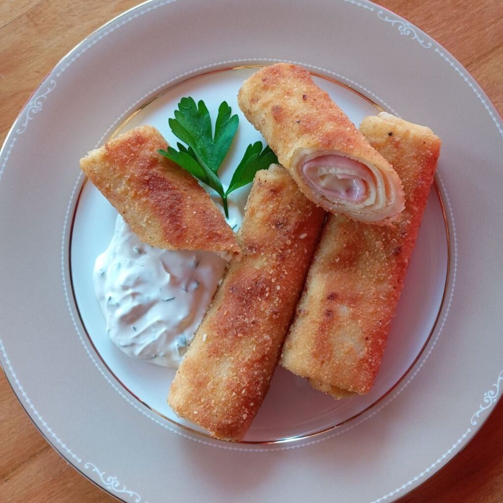 Three Crispy Ham Crepe Rolls served with creamy sauce and parsley garnish, one cut in half to show the filling.