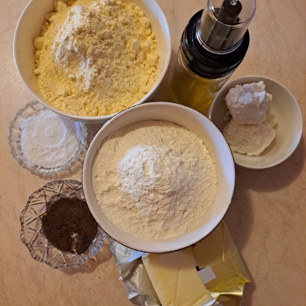 Ingredients for Cornmeal Tartlets on a kitchen table: cornmeal, flour, salt, black pepper, olive oil, butter, and goat cheese.